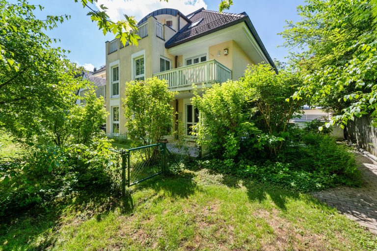 Ein mehrstöckiges Haus mit einem Balkon und einem grünen Gartenbereich umgeben von Bäumen.