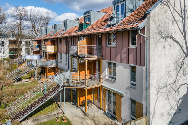 Mehrfamilienhaus mit mehreren Balkonen und Treppen im Freien, umgeben von Bäumen.