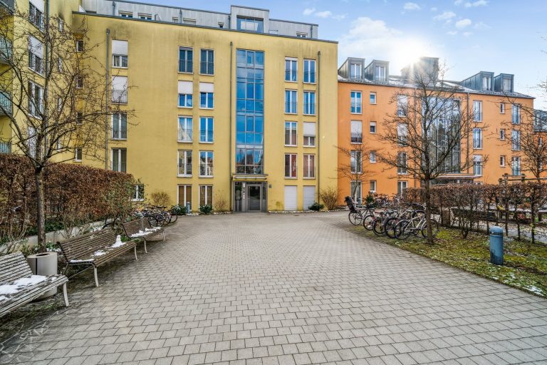 Eingang zu einem Wohngebäude mit gelber und orangefarbener Fassade und gepflastertem Vorplatz.