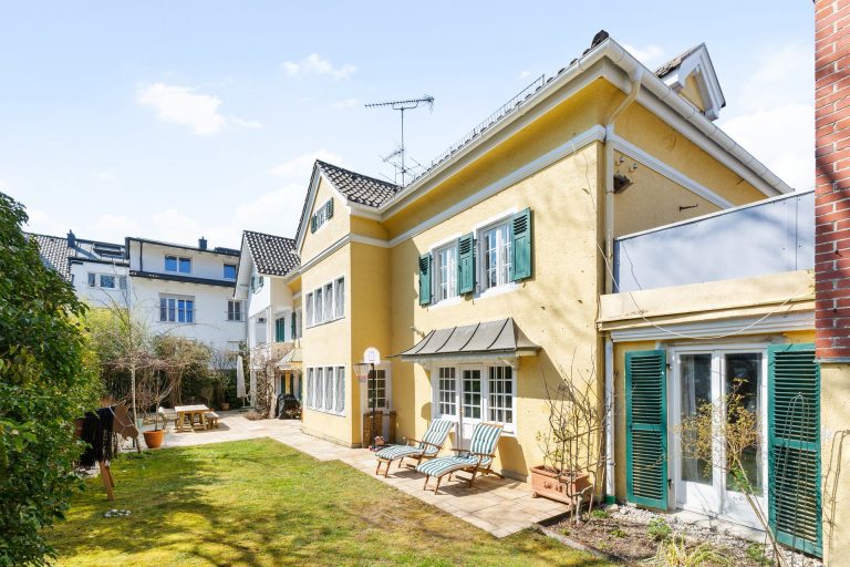 Gelbes Einfamilienhaus mit grünen Fensterläden und Gartenmöbeln im Freien.
