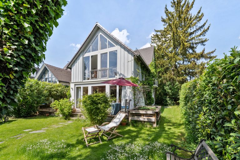 Ein modernes Haus mit großen Fenstern, einem Garten und einer Terrasse mit Sonnenschirm.