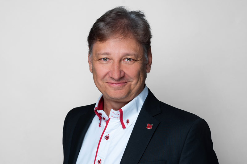 Mann mit grauen Haaren, Anzug und Hemd mit roten Akzenten, lächelt in die Kamera.