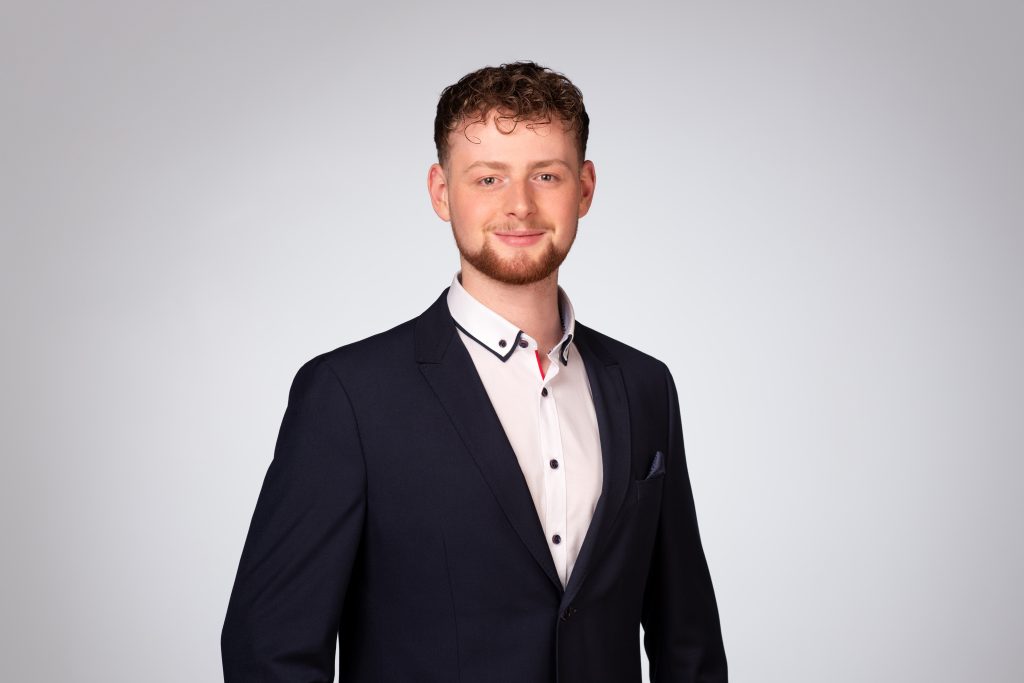 Mann mit lockigem Haar trägt einen dunklen Anzug und ein weißes Hemd in einem neutralen Hintergrund.