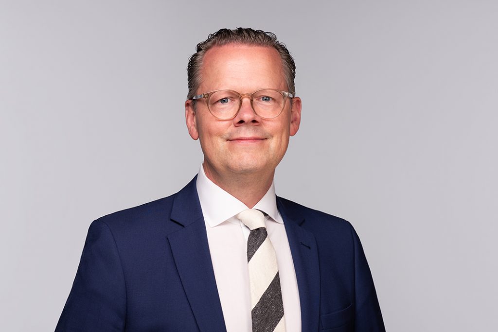 Mann mit Brille und Anzug, der in die Kamera lächelt, vor neutralem Hintergrund.