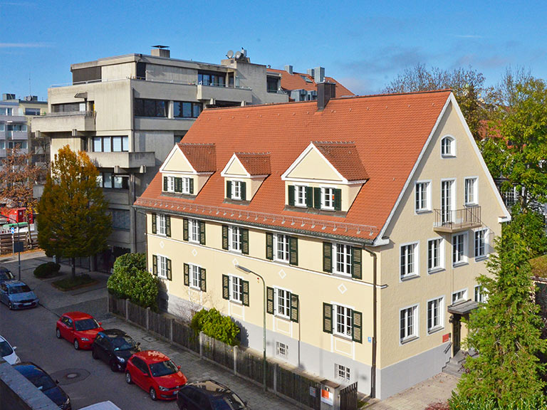 Mehrfamilienhaus mit rotem Satteldach, grünen Fensterläden und mehreren Fenstern in einer städtischen Umgebung.