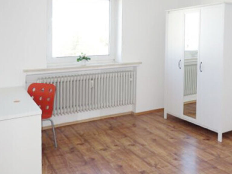 Ein leeres Zimmer mit einem weißen Schreibtisch, einem roten Stuhl und einem Kleiderschrank mit Spiegel.