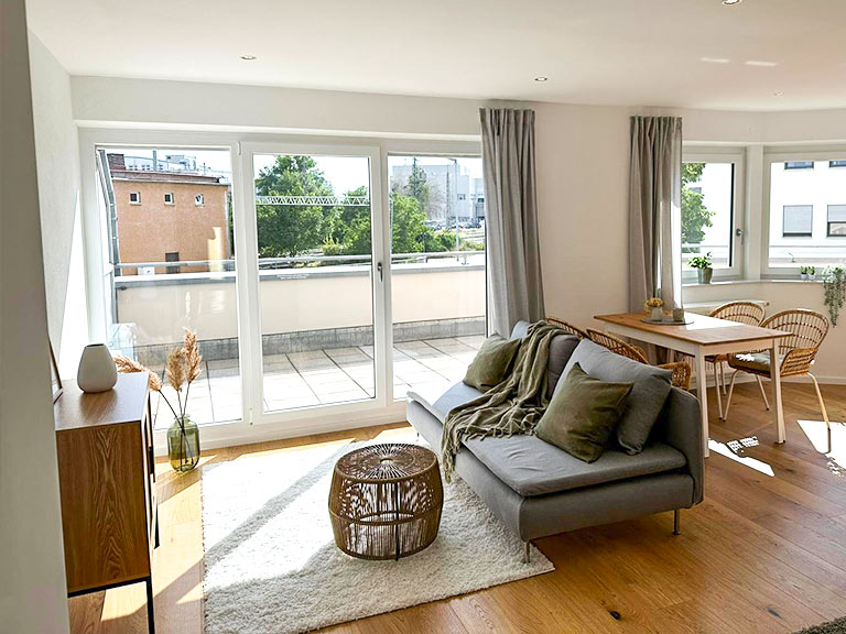 Helles Wohnzimmer mit Sofa, Tisch und großen Fenstern, die auf einen Balkon blicken.