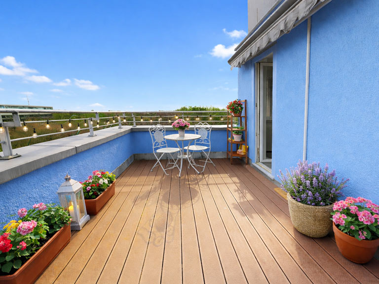 Terrassenblick mit Holzterrasse, blauem Pool und Sitzbereich mit Metallmöbeln, dazu Blumen in Kübeln und Balkonpflanzkästen vor blauem Himmel