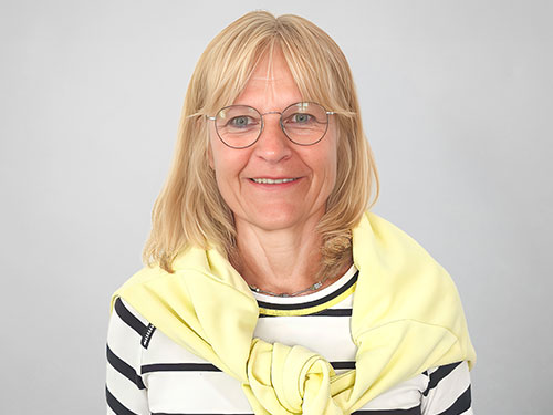 Claudia Netzer mit blonden Haaren und Brille trägt ein helles Tuch vor neutralem grauem Hintergrund.
